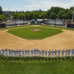 Elevated drone shot of the 2023 Redding Colt 45s team in a single line, offering various gestures toward the camera, in the grass along the base path, behind second base, with the pitcher's mound, home plate and the stands in the background.